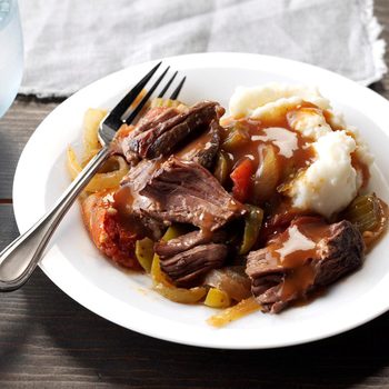 Plate holds pot roast with vegetables and mashed potatoes, drizzled with gravy. A fork rests on the side, atop a wooden table.