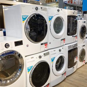 Washing machines stacked on display with sale tags in a brightly lit appliance store aisle.