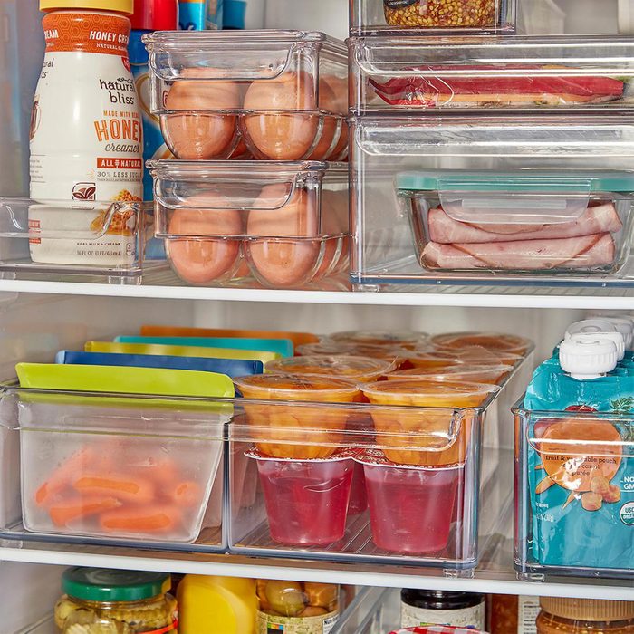 Refrigerator shelves store eggs, juice, meats, and containers in clear bins, organized with creams and pickles. Packages of sliced fruit and snacks are visible.