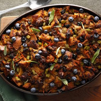 Casserole sits on a green napkin, filled with toasted bread, blueberries, and herbs, resting on a wooden tabletop with silverware nearby.
