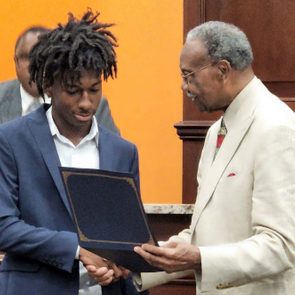 Two men examine a certificate; one hands it to the other in a formal indoor setting.