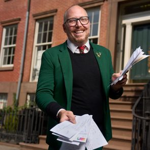 A person smiles while handing out envelopes in front of a brick building with large windows and a staircase.