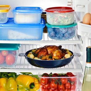 Roast chicken sits on a refrigerator shelf surrounded by assorted vegetables, plastic containers, eggs, and bottles, creating a well-stocked and organized fridge scene.