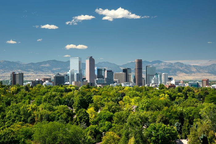 Skyscrapers stand tall against a mountain backdrop, surrounded by lush green trees under a clear blue sky with scattered clouds.