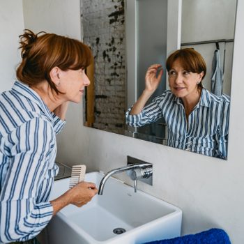 Woman examines face in bathroom mirror, holding a comb, surrounded by a white sink and blue towel, wearing a striped shirt.
