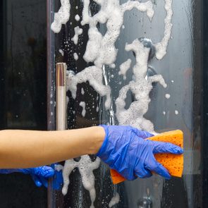 Hand wearing blue glove cleans shower glass with sponge covered in soap suds. Showerhead visible in the background.