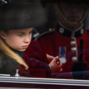 A child, wearing a black hat, sits inside a vehicle, next to a uniformed person, visible through the window.