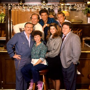 A group of six people pose cheerfully in a bar setting, surrounded by glasses and wooden decor.