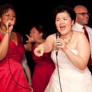 Two women sing passionately into microphones; one wears a white dress, the other a red dress, with laughing people behind them at an event.