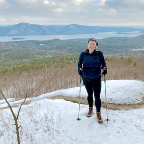 Hiker stands on snowy trail, holding trekking poles, with mountains and a lake in the background under a cloudy sky.