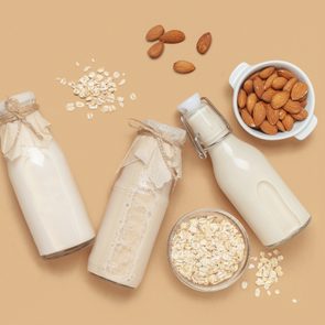 Bottles of milk lie with almonds and oats scattered around, on a beige background, alongside small bowls of whole almonds and oats.