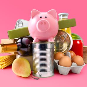 Piggy bank sits atop a tin can surrounded by groceries, including eggs, pasta, a pear, and canned goods, against a pink background.