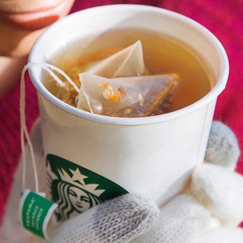 A gloved hand holds a Starbucks cup with pyramid tea bags. The person wears a red sweater in a cozy outdoor setting.