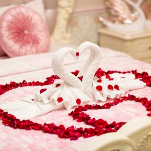 Two towel swans form a heart, surrounded by red rose petals, on a pink bed with decorative pillows.
