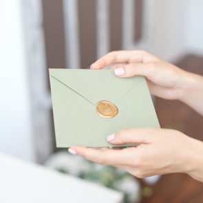 Hands hold a green envelope sealed with a gold wax stamp; the background is softly blurred.