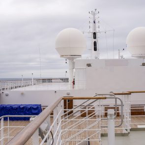 Satellite domes stand stationary on a cruise ship deck with wooden railings, surrounded by a cloudy sky and ocean.