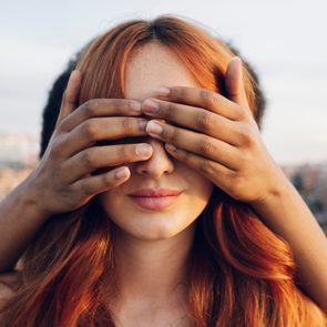 Hands cover a woman's eyes, set against a blurred cityscape background.