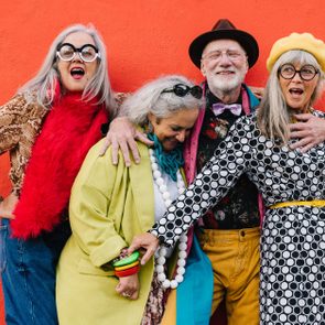 Four joyful people with vibrant, eclectic outfits pose against a vivid orange wall, expressing happiness and camaraderie.