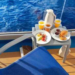 Breakfast on table, with pastries and fruit beside ocean-view window on cruise ship deck, surrounded by blue waters and a blue chair.