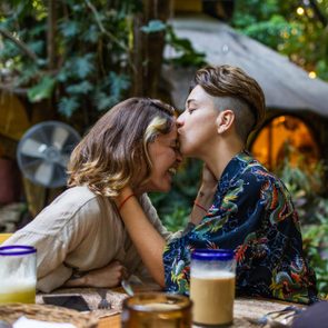Two people embrace, one kissing the other's forehead, seated at a table outdoors surrounded by greenery and colorful lights.