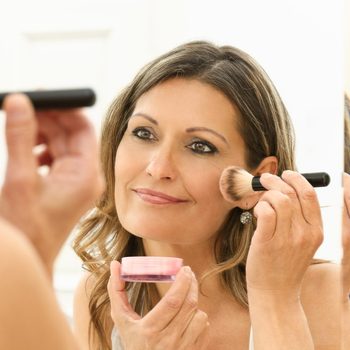 Woman applies makeup with a brush while looking in a mirror, holding a small container. Bright, indoor setting.