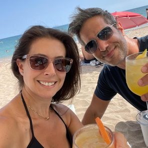 Two people smile, holding drinks on a sandy beach with the ocean and a red umbrella in the background.