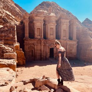 A person stands on a rock, overlooking Petra's ancient stone facade, with rocky cliffs under a clear blue sky.