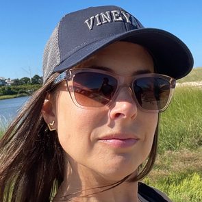 Woman wearing sunglasses and a "VINEYARD" cap, standing by a grassy riverbank on a sunny day.