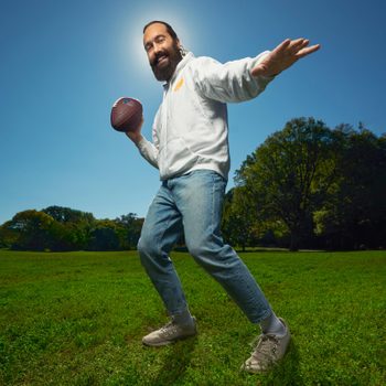 Man holding football, preparing to throw, stands on vibrant green grass under clear blue sky, with trees in the background.