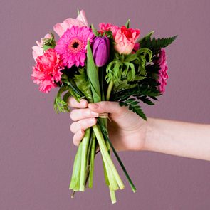 A hand holds a vibrant floral bouquet against a plain mauve background.