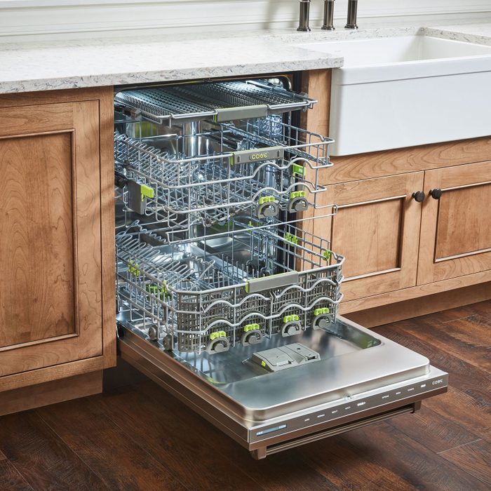 Dishwasher with pulled-out racks, situated in a kitchen with wooden cabinetry and a marble countertop, standing next to a white farmhouse-style sink.