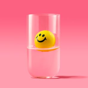 A yellow smiley face ball floats in a glass of water against a pink gradient background.
