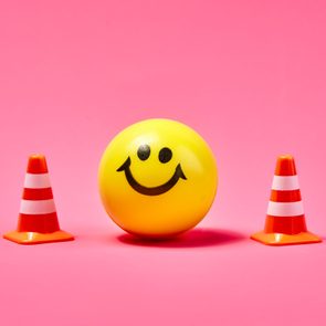 A yellow smiley face ball is placed between two small orange traffic cones on a solid pink background.