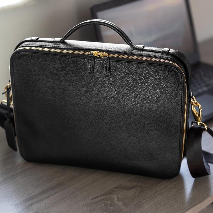 Black leather briefcase rests on a wooden desk, with a laptop in the background, under soft lighting.