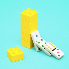 Dominoes lean against a yellow Lego block stack on a turquoise background.