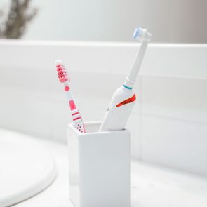 Toothbrushes stand upright in a white holder on a bathroom counter near a faucet, with blurred plants in the background.