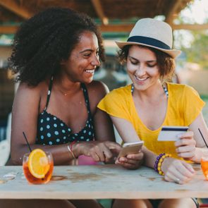 Two people smile while looking at a phone; one holds a credit card. They're at a table with drinks, outdoors, under a wooden roof.