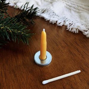 Small yellow candle stands on a wooden table with a matchstick and pine branch nearby.
