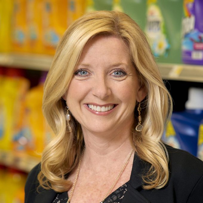 A person smiling stands in front of colorful detergent bottles on grocery store shelves.