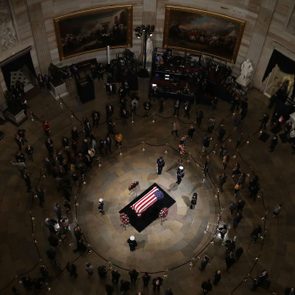Coffin displays U.S. flag, surrounded by mourners and guards, in a grand circular hall adorned with large paintings and statues.