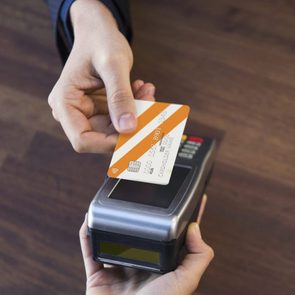 A hand taps an orange card on a payment terminal, held by another hand, against a wooden background.