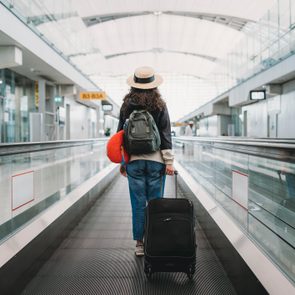 Traveler walks with suitcase on airport moving walkway, wearing a hat and carrying a backpack. Signs display: 