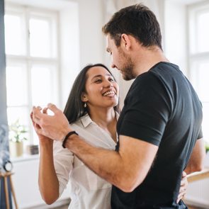 Two people smiling and dancing in a bright living room with framed pictures on the wall and large windows letting in sunlight.