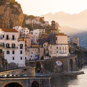Buildings perch on rocky cliffs, overlooking a calm sea at sunset with mountains in the background.
