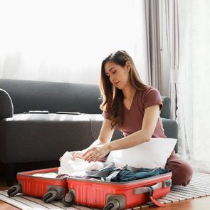 Woman packs clothes into red suitcase, sitting on a carpeted floor in a sunlit living room with a sofa and indoor plant nearby.