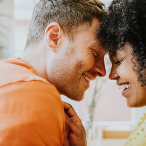 Two people smiling, touching foreheads, in a bright room with plants in the background.