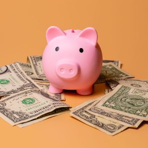 Pink piggy bank sits surrounded by scattered dollar bills on an orange background.