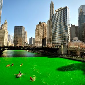 Kayakers paddle on a bright green river, surrounded by towering skyscrapers and a crowded bridge under a clear sky.