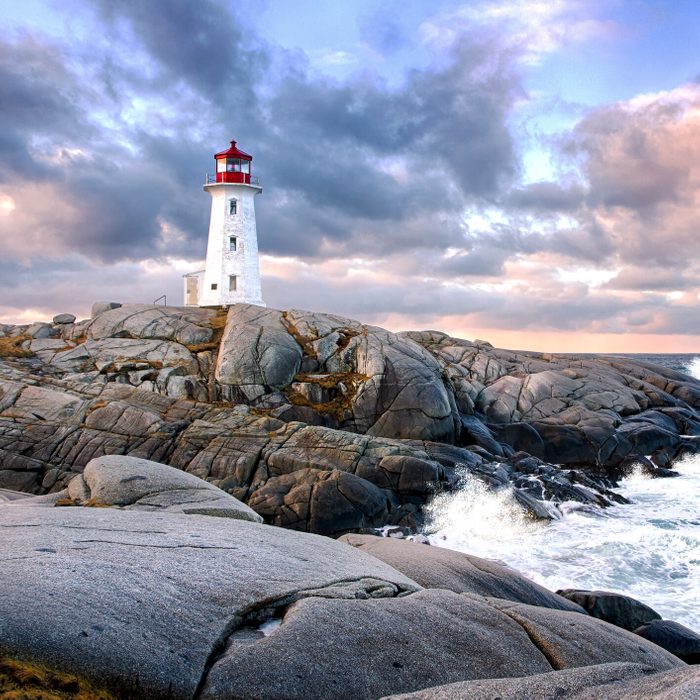 A lighthouse stands on rocky cliffs, overlooking ocean waves under a dramatic, cloudy sky at sunset.