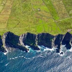 Steep green cliffs descend sharply into the crashing ocean waves, forming a dramatic coastal landscape under a bright blue sky.